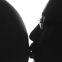 Eine stimmungsvolle Schwarz-Weiß-Gegenlichtaufnahme, die die Silhouette eines Mannes zeigt, der den Bauch einer schwangeren Frau küsst. Ein intimer und emotionaler Moment der Vorfreude, meisterhaft festgehalten von Fotograf Michael Schalansky.